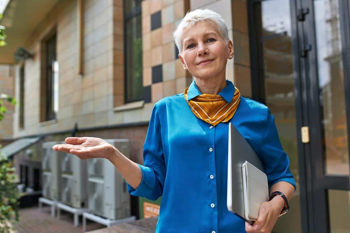 Stylish confident middle aged businesswoman with short hairstyle posing outside office building with laptop under her arm, making gesture as if holding something on hand. People, age, work and gadgets